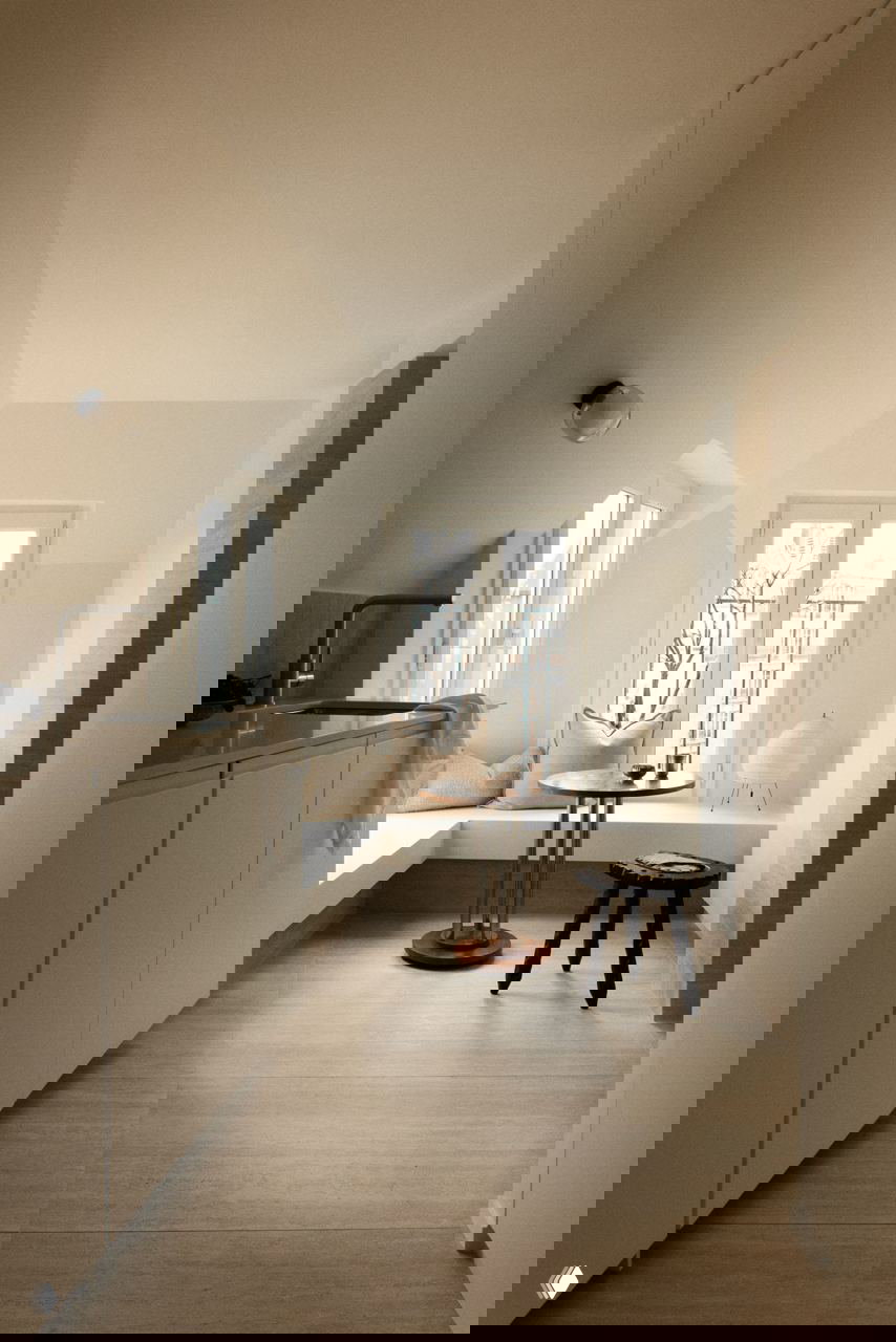 Кухонный уголок у окна: встроенные белые шкафы, скамья с подушками, небольшой круглый стол и мягкое естественное освещение из окон.