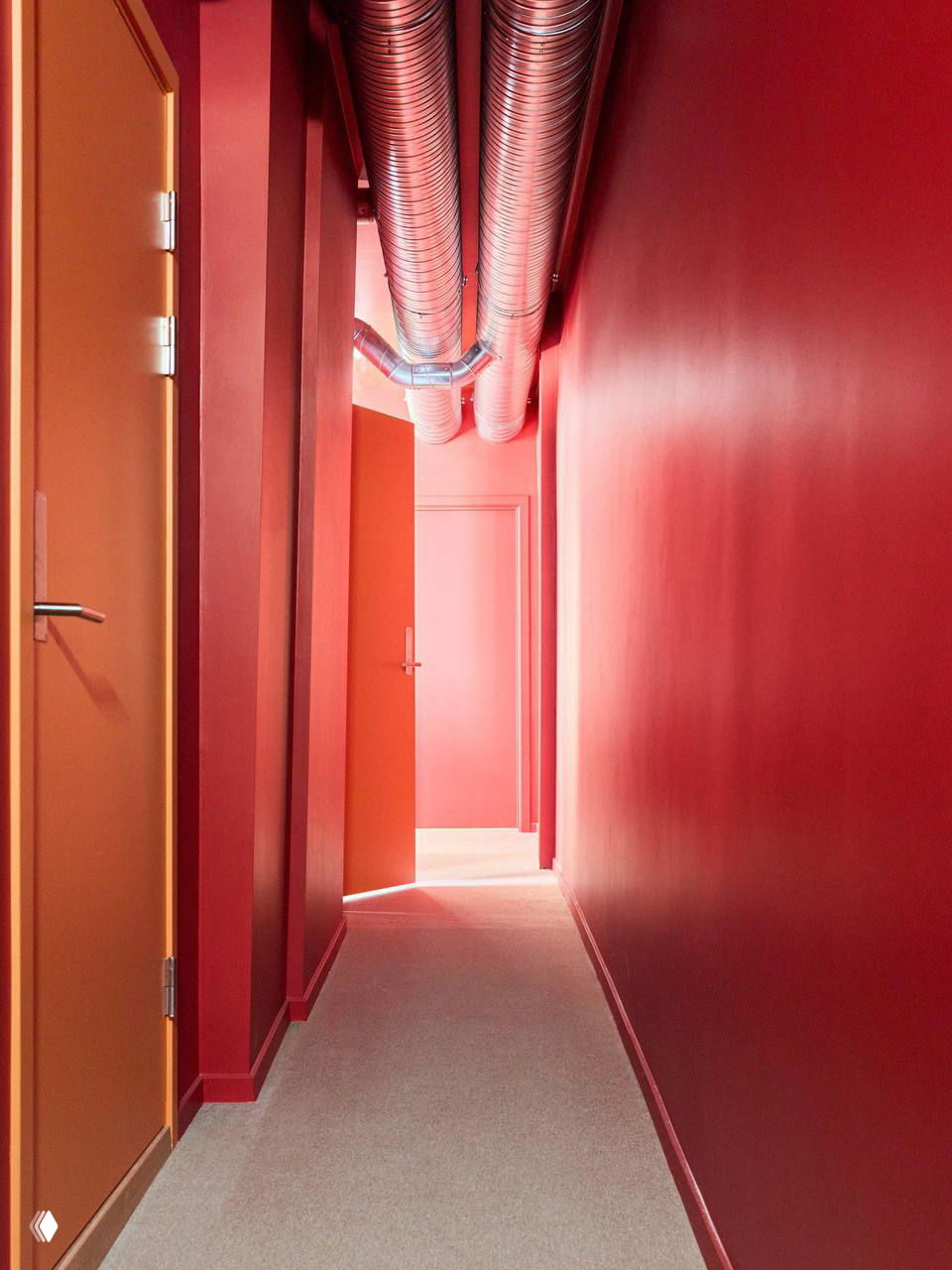 Яркий коридор студии в насыщенных красно-оранжевых тонах: однотонные стены и двери, открывающие доступ в помещение с низкими воздуховодами на потолке.