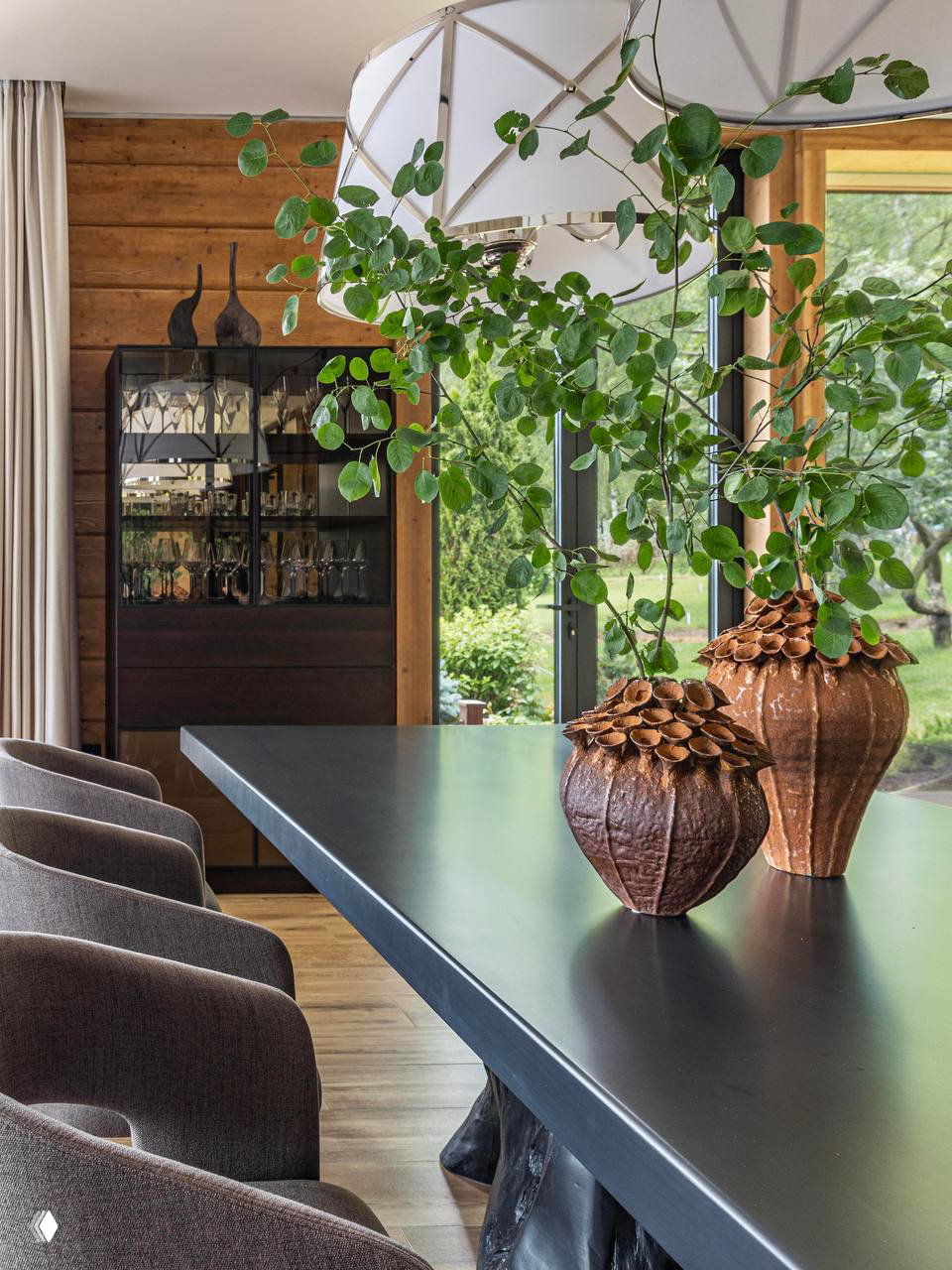 Столовая зона с чёрным столом и крупной подвесной люстрой; декоративные вазы и деревянная стена создают тёплую, естественную атмосферу интерьера.