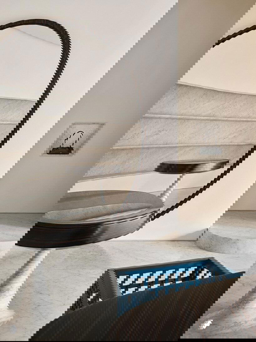 Интерьерный кадр с большим круглым зеркалом, мраморными столами и клетчатым синим креслом как элементом скульптурности и контраста в пространстве.