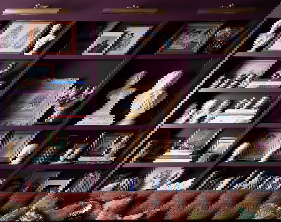 Тёмно-фиолетовая полка с бюстами, картинами и рамками, множество маленьких антикварных предметов и ретро-декор на фоне текстурированного дивана.