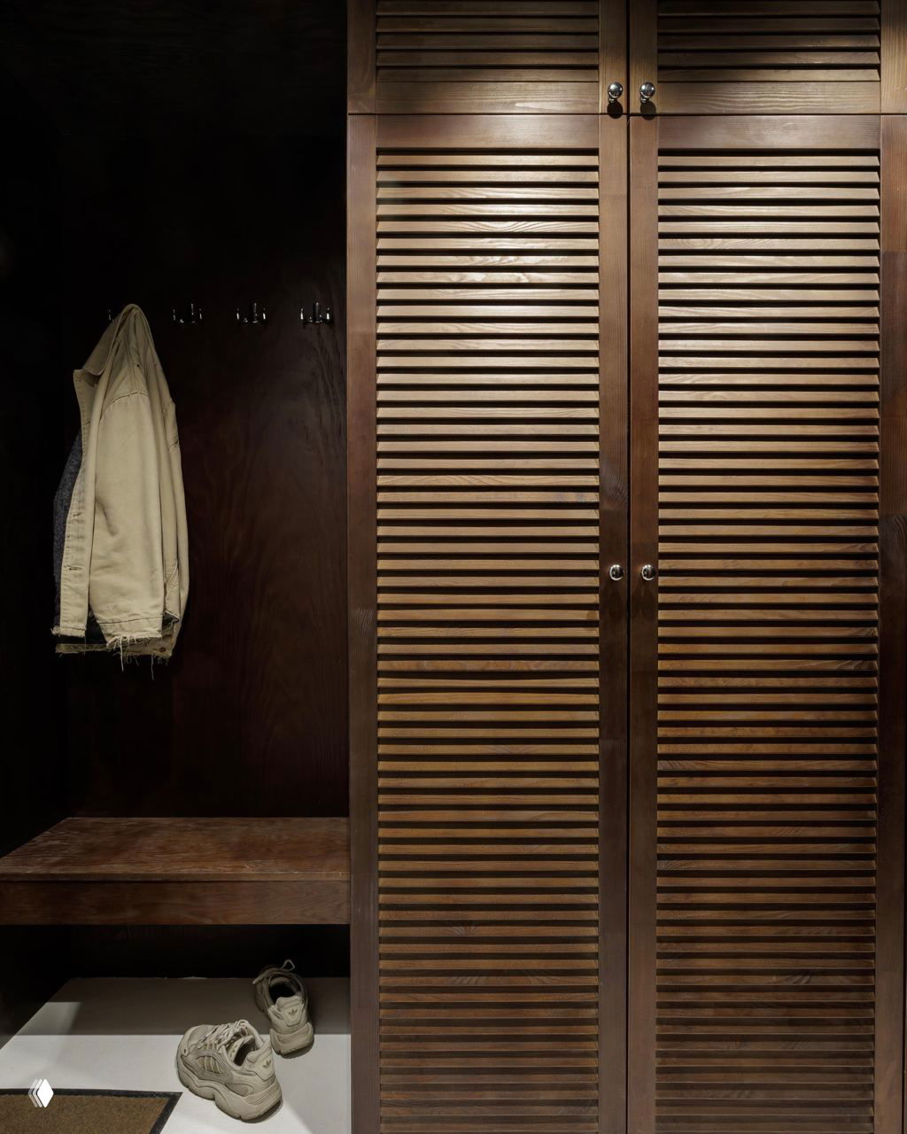 Прихожая со стеллажом и деревянными жалюзийными дверцами шкафа, тёмное оформление и аккуратно размещённая обувь у входа
