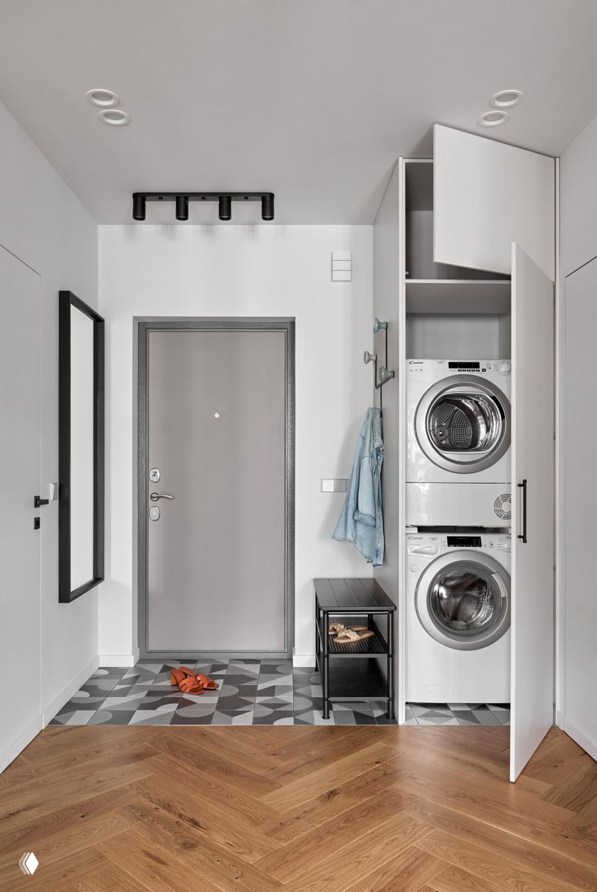 Прихожая с открытым встроенным шкафом, где видны стиральная и сушильная машины, черная мебельная тумба и рисунок напольной плитки у двери.