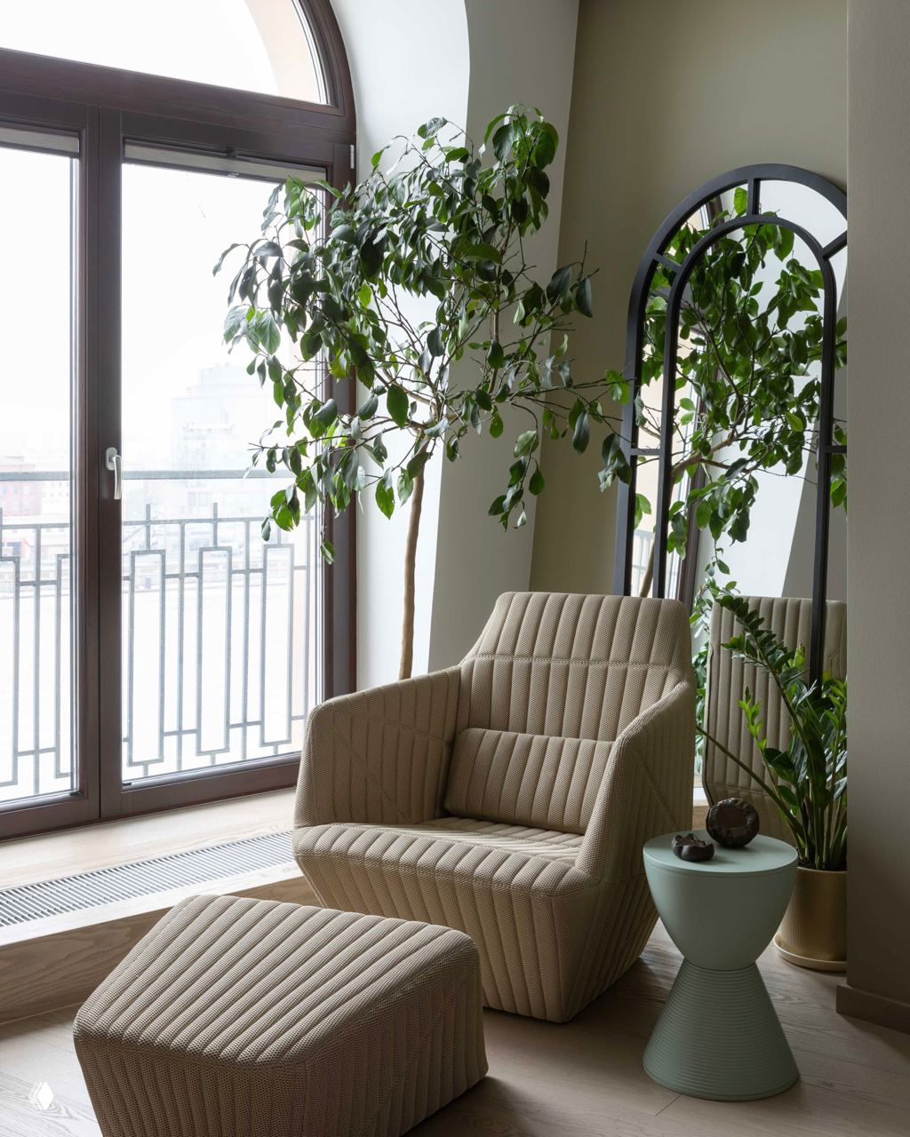 Уютный угол с креслом у панорамного окна и высоким фикусом или крупным лиственным растением рядом с зеркалом, создающим глубину интерьера.