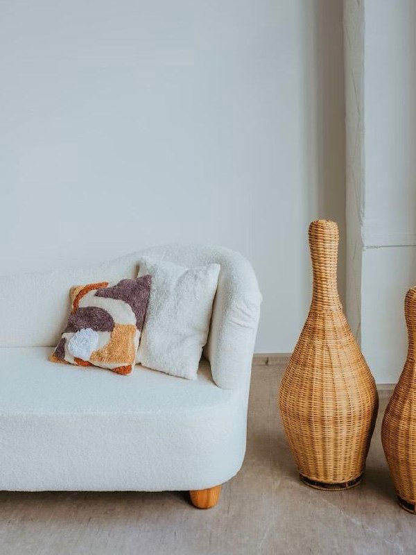 Белый изогнутый диван в стиле букле рядом с высокой плетёной вазой и декоративными подушками на нейтральном полу — уютный уголок скандинавского вдохновения.