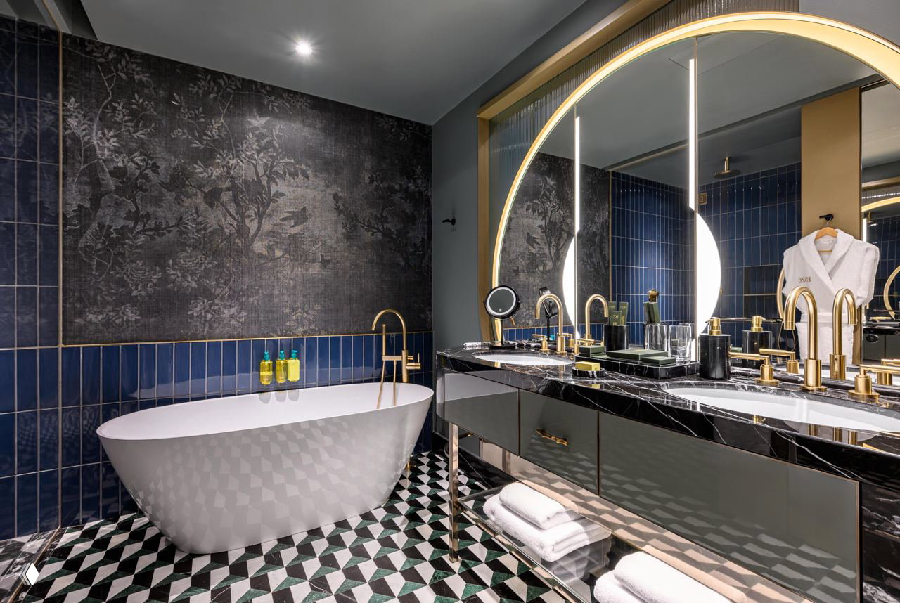 Люксовая ванная номера Enzo Hotel: свободностоящая ванна, чёрно-белая мраморная столешница с золотой фурнитурой и декоративная узорчатая плитка на полу.