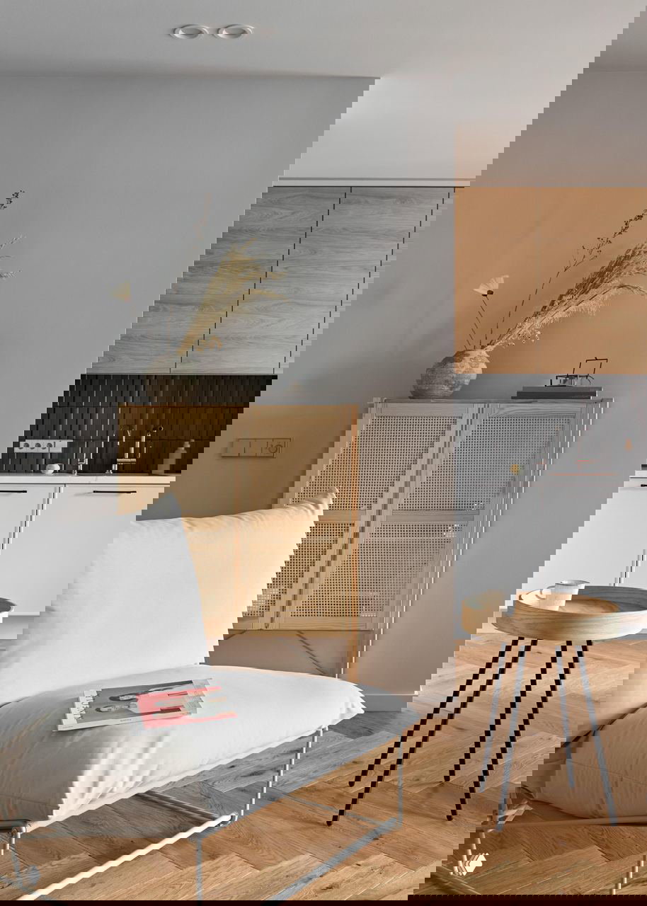 Деталь интерьера: ротанговая тумба с вазой и сухими травами, уютное белое кресло и часть кухни с деревянными фасадами на нейтральном фоне.