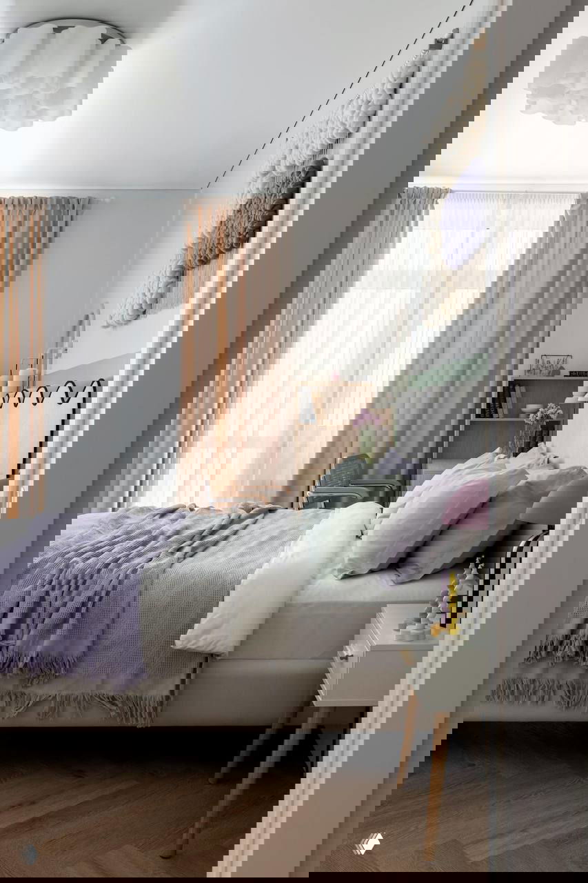 Спальня — вид с изголовья: сиреневое покрывало, светло-зеленые подушки и тумбочка с яркой настольной лампой и свечами.