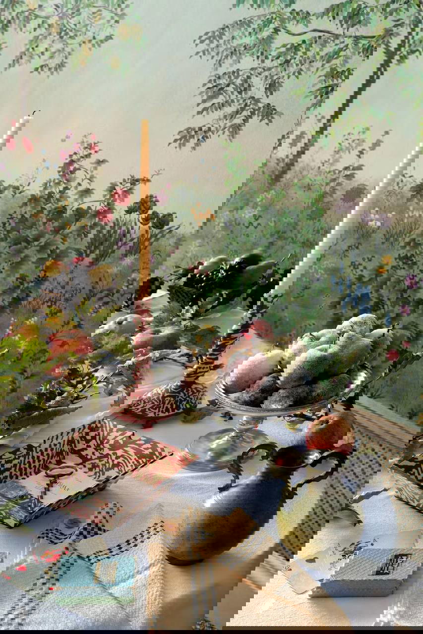 Крупный план столика с фруктами, высокой свечой и декоративными предметами на фоне обоев Fruit Garden с изображением деревьев и полевых цветов.