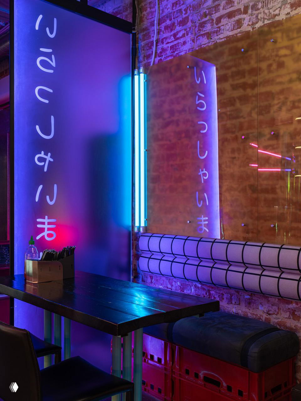 Столик у матовой перегородки с японскими символами и голубо‑фиолетовой неоновой подсветкой, скамья и кирпичная стена на заднем плане.