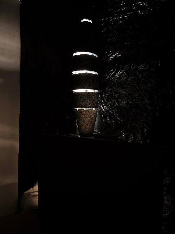 Тёмная инсталляция с вертикальным подсвеченным объектом: игра света и фактуры, создающая сакральную или медитативную атмосферу в экспозиции.