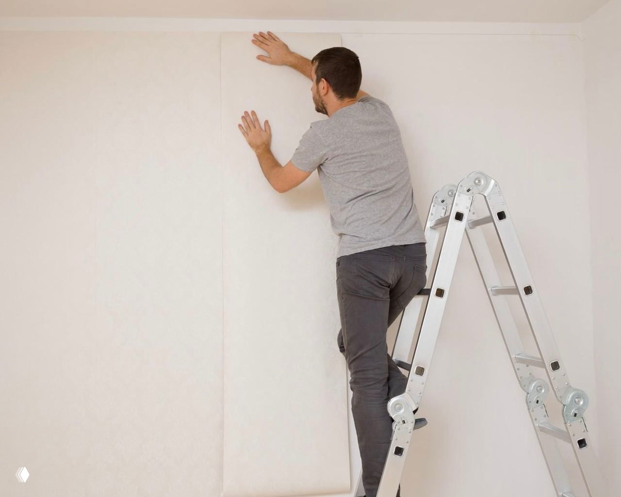 Человек на стремянке клеит светлые полотна обоев на стену; видна работа по выравниванию и сглаживанию поверхности при помощи флизелиновых материалов.