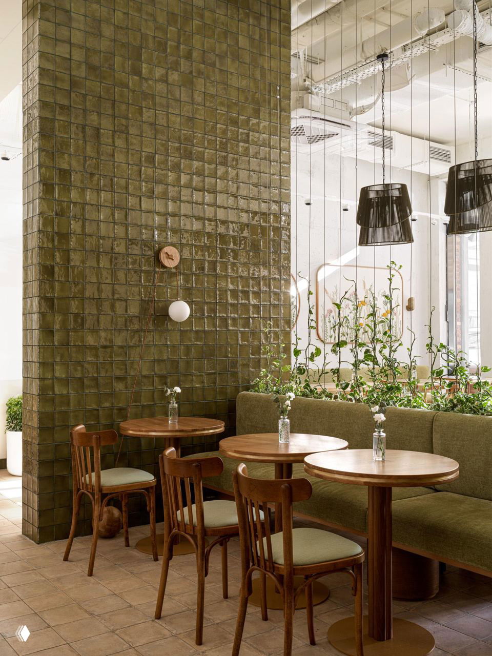Уютный уголок кафе: деревянные столы, мягкий зелёный диван и плетёная люстра на фоне декоративной плитки и живых растений в интерьере.