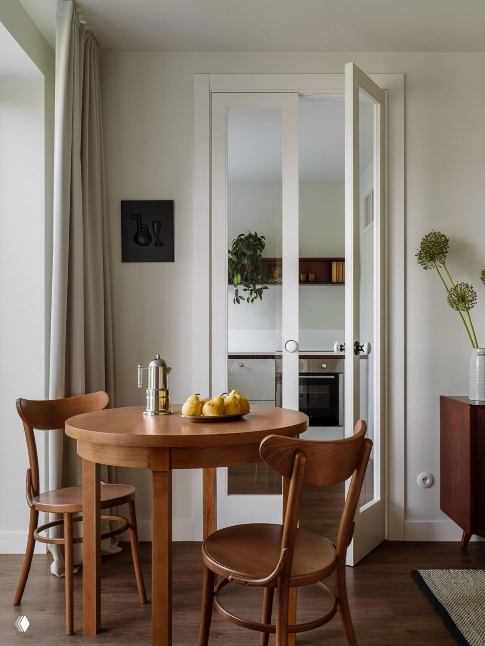 Обеденная зона: круглый деревянный стол и винтажные стулья у высоких дверей, ведущих в кухню, естественное освещение и простая стилистика.