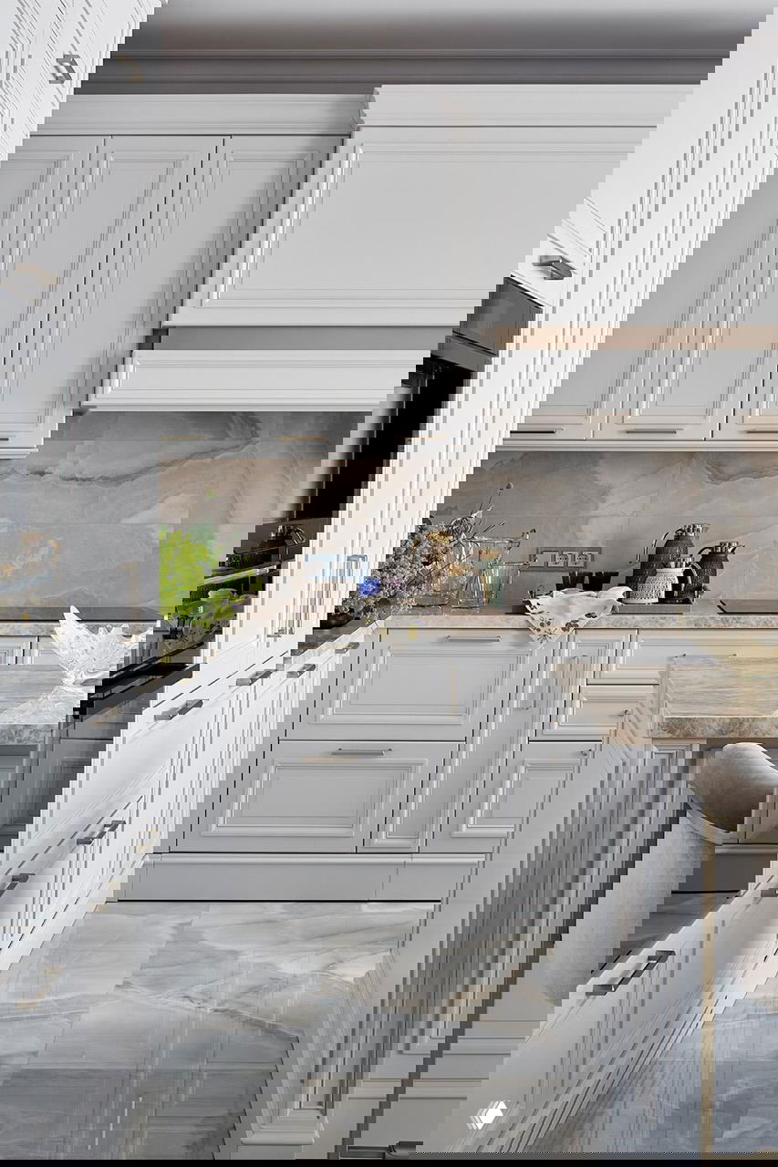 Кухонная зона с белыми резными фасадами, мраморной столешницей и фартуком, барные стулья с обивкой и аккуратные латунные ручки на шкафах.