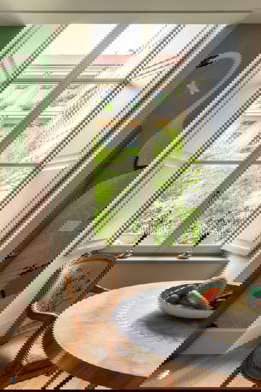 Окно с распахнутыми ставнями: столовая зона с круглым столом и стулом в классическом стиле, вид на балкон и зелёные растения.