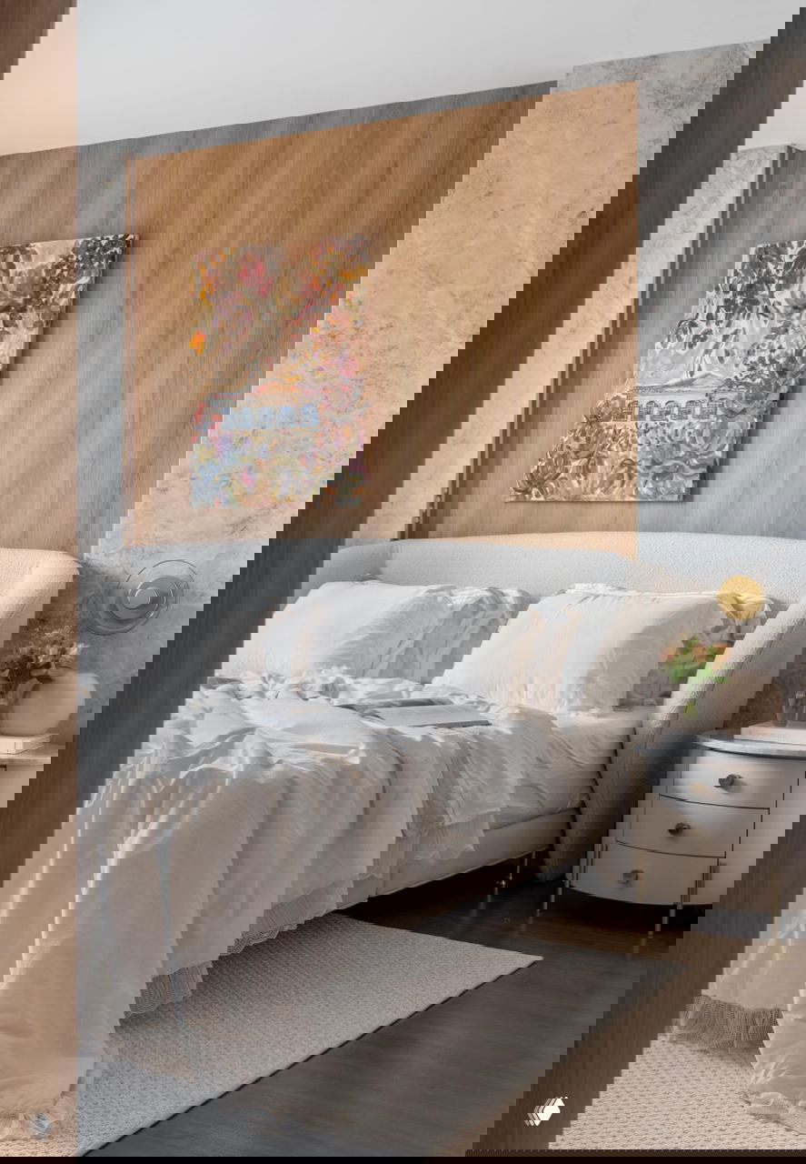 Спальня с мягкой белой кроватью, обложенной текстилем, деревянной панелью за изголовьем и живописной картиной на стене над кроватью.