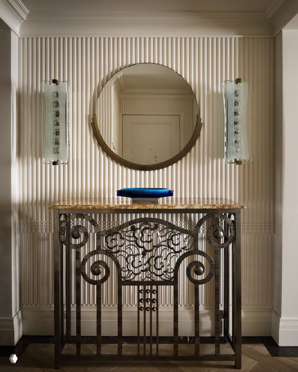 Декоративная консоль в ар‑деко стиле с витой металлической решёткой, круговым зеркалом и рельефной панелью на стене в коридоре квартиры.