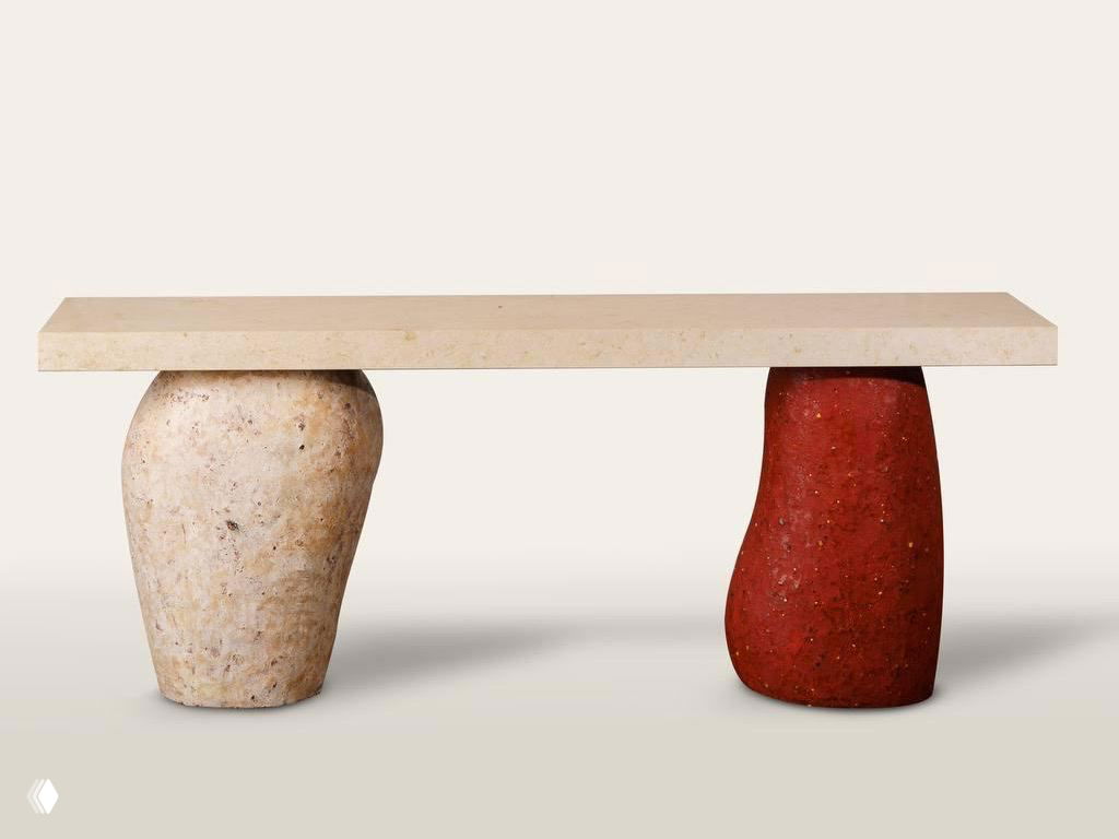 Длинный стол с массивной столешницей и двумя скульптурными опорами из переработанного материала: одна светлая, другая тёпло-красная, минималистичная презентация изделия.