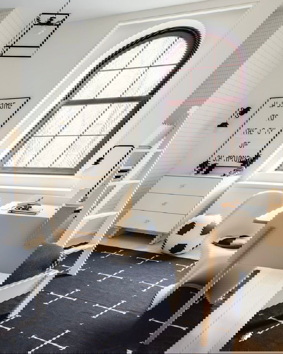 Светлая комната с арочным окном: письменный стол, комод, мягкий ковёр и футбольный мяч — лаконичный и функциональный интерьер для мальчика.