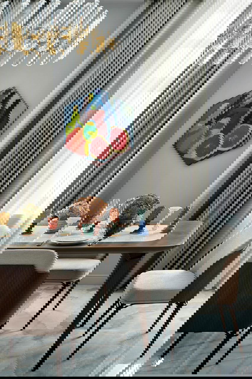 Столовая зона с деревянным столом, разноцветными стеклянными предметами и независимой картиной на фоне серо-голубой палитры интерьера, подчёркнутой латунными деталями.