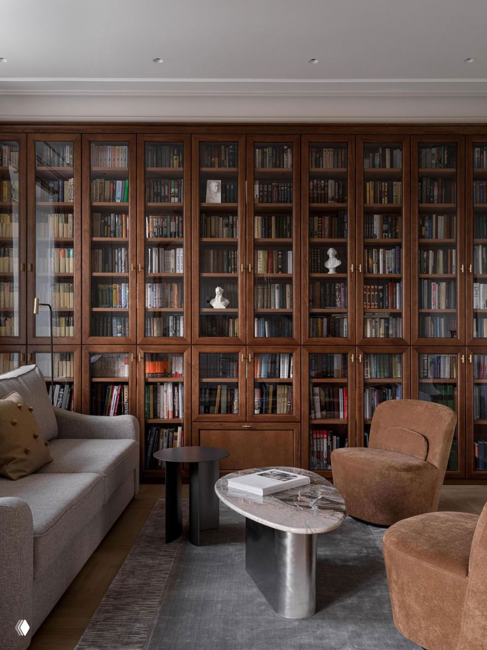 Гостиная с большой книжной стеной в тёплом деревянном тоне: стеклянные дверцы шкафов, кожаные и тканевые кресла, низкие столики и мраморная столешница.