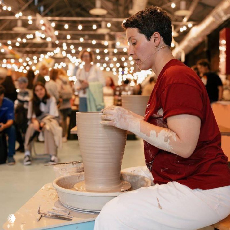 Фотография с визуальными артефактами: мастер за гончарным кругом и посетители на фоне праздничных гирлянд, кадр выглядит повреждённым цифровыми искажениями.