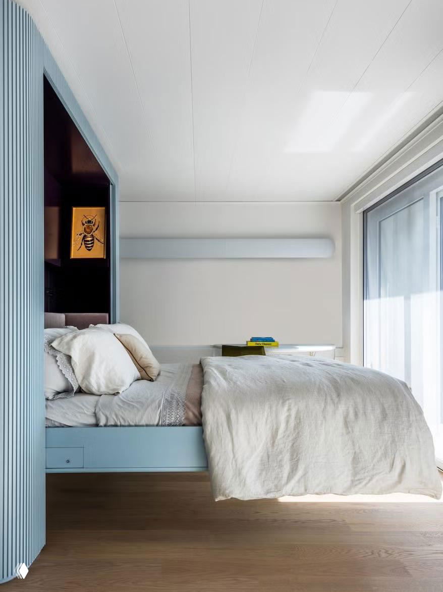 Пастельная светлая спальня с полукруглой голубой перегородкой вокруг кровати и большим окном — минималистичное разделение пространства без полноценных стен.