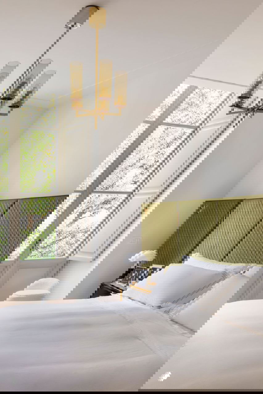 Спальня с высоким окном и зелёным текстильным изголовьем, белое постельное бельё и минималистичная декорация в спокойных тонах.