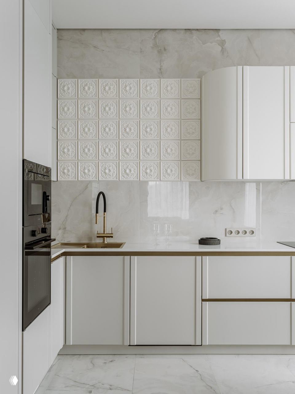 Крупный план кухни: светлый каменный фартук, декоративные рельефные плитки и белые радиусные фасады в сочетании с латунным смесителем