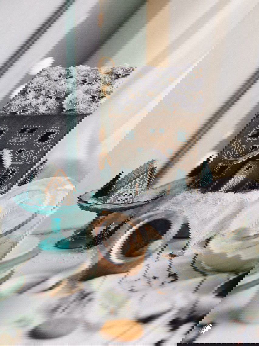 Имбирный пряничный домик с «снежной» глазурью и миниатюрными декорациями вокруг, снятый в тёплом светлой атмосфере кухни.