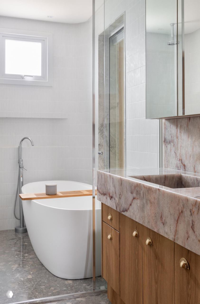 Ванная комната с отдельно стоящей белой ванной, мраморной столешницей и деревянной мебелью под раковиной, светлый современный интерьер.