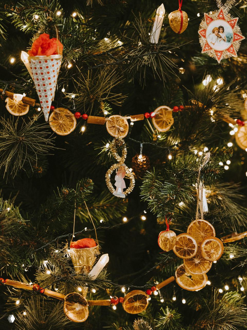 Ёлка, украшенная гирляндой из сушёных апельсиновых кружочков, палочек корицы и небольших украшений; тёплая гирляндная подсветка создаёт уютную атмосферу.
