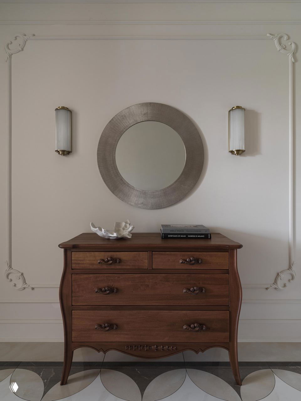 Прихожая с деревянным комодом в классическом стиле, круглоe зеркало и элегантная лепнина на пастельно-бежевой стене, пол с декоративной мозаикой.