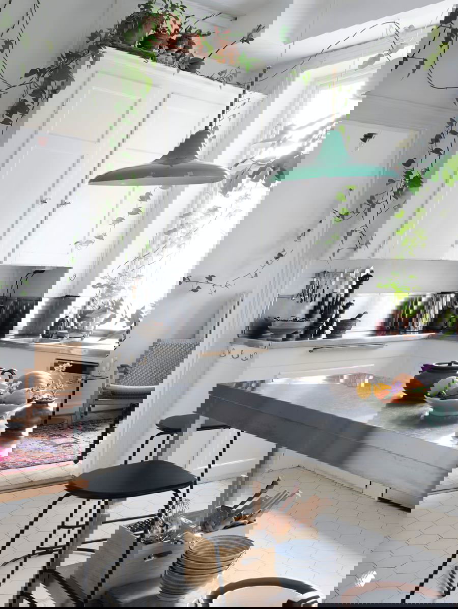 Светлая кухня с белыми фасадами и зелёными лианами, спускающимися по шкафам; металлизированная рабочая поверхность и простой стол.