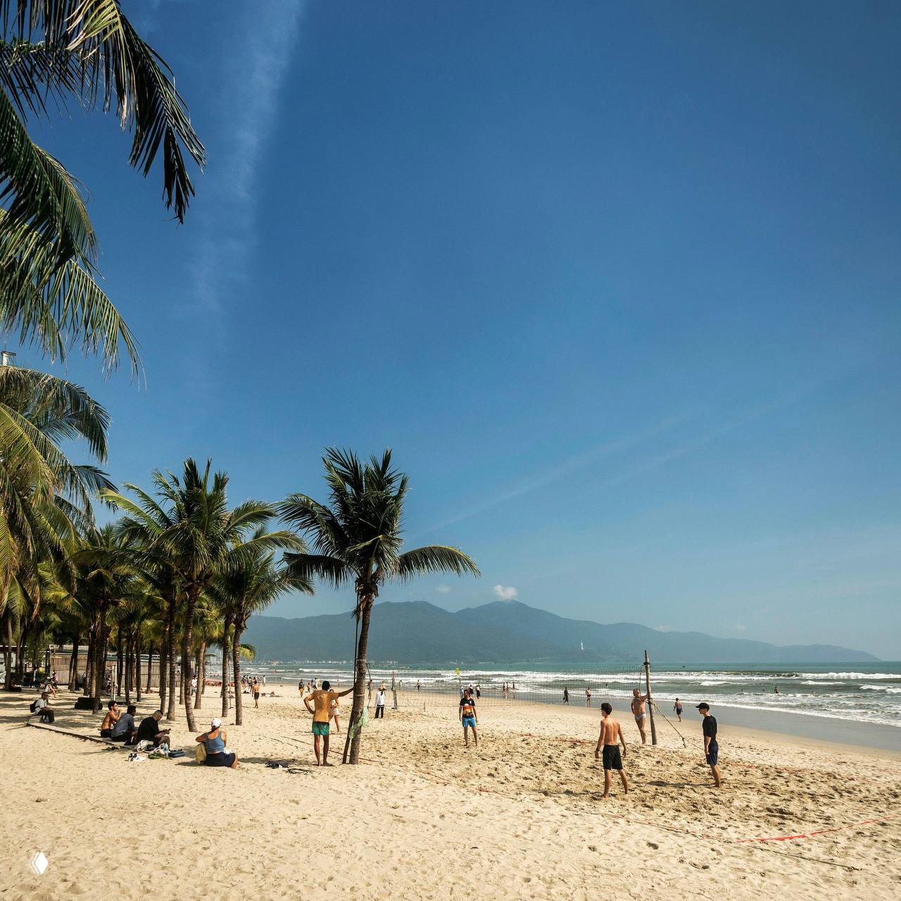 Песчаный пляж Дананга с пальмами на берегу и людьми, отдыхающими и играющими в волейбол на фоне гор и моря под ясным небом.