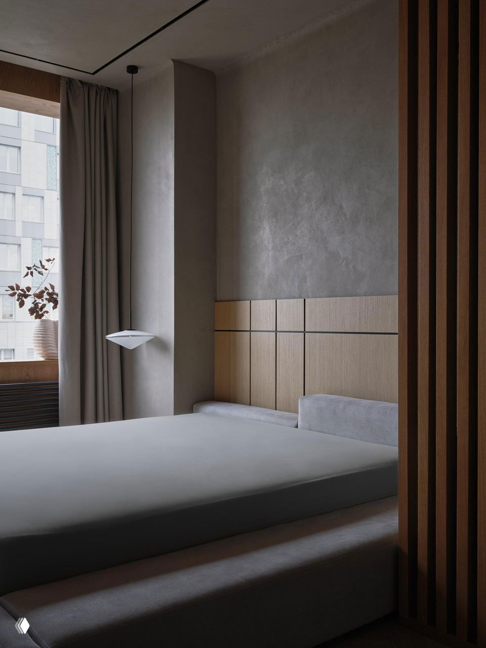 Спальня в спокойных нейтральных тонах: минималистичная низкая кровать, серая текстурная штукатурка на стене, деревянные панели и мягкое дневное освещение без лишнего декора.