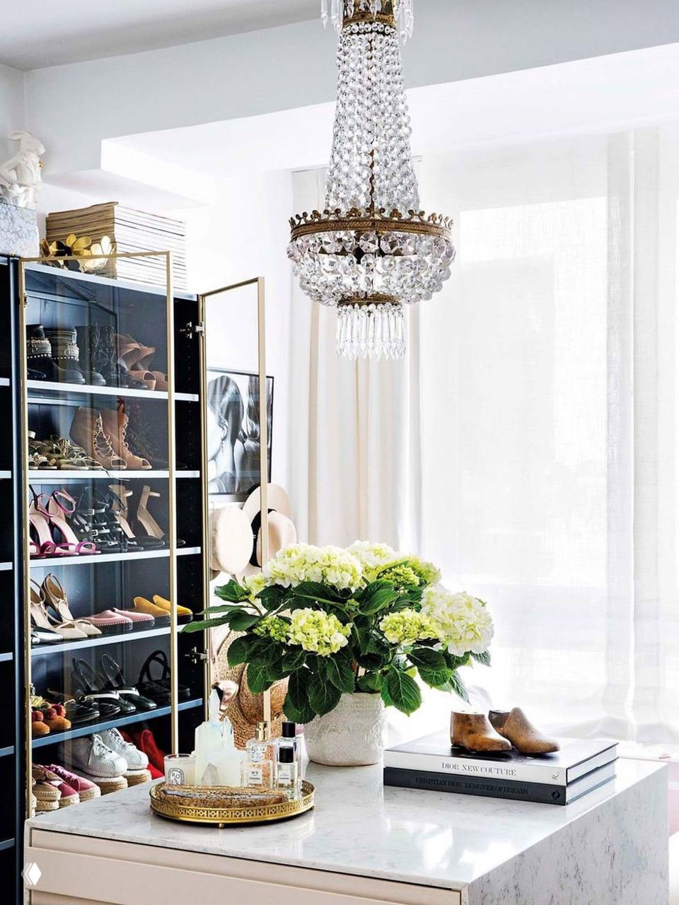 Гардеробная с прозрачным шкафом для обуви, мраморной столешницей и вазой с гортензиями на островке — светлый, организованный уголок.