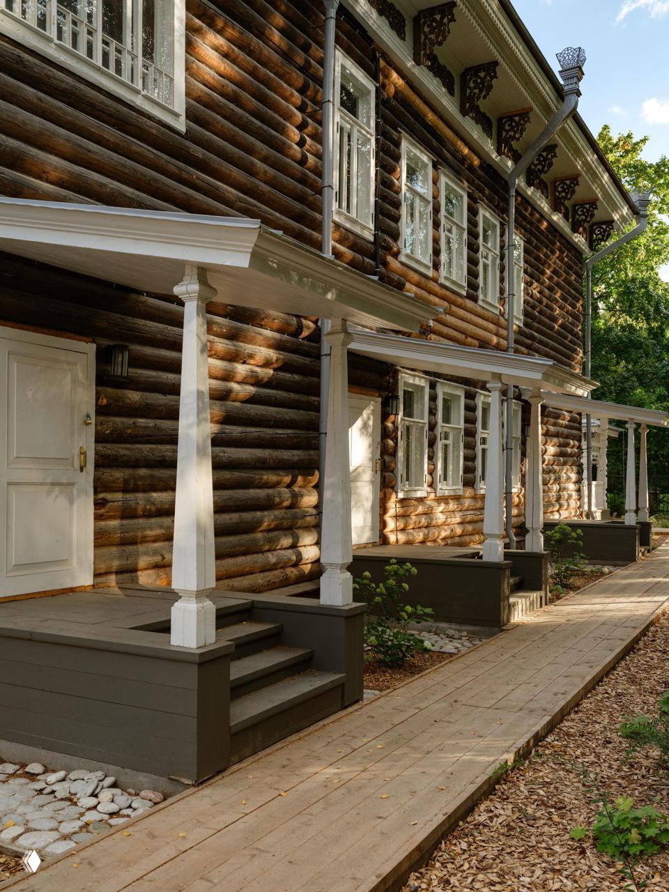 Бревенчатый фасад доходного дома 1915 года с верандами и колоннами, деревянная отделка и тропинка вдоль здания в зелёной обстановке.