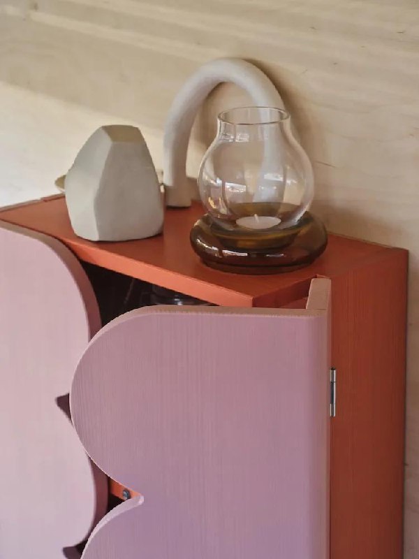Кадр с деталями интерьера: розовая мебельная дверца с округлым краем, на поверхности — стеклянный подсвечник и декоративные керамические объекты.