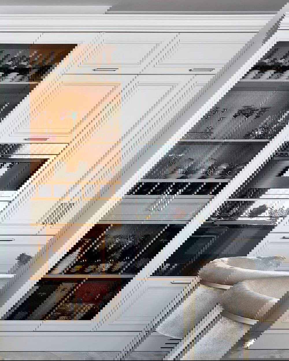 Белая корпусная мебель с витриной и деревянными внутренними полками, где аккуратно размещены бутылки и посуда в стиле современной классики.