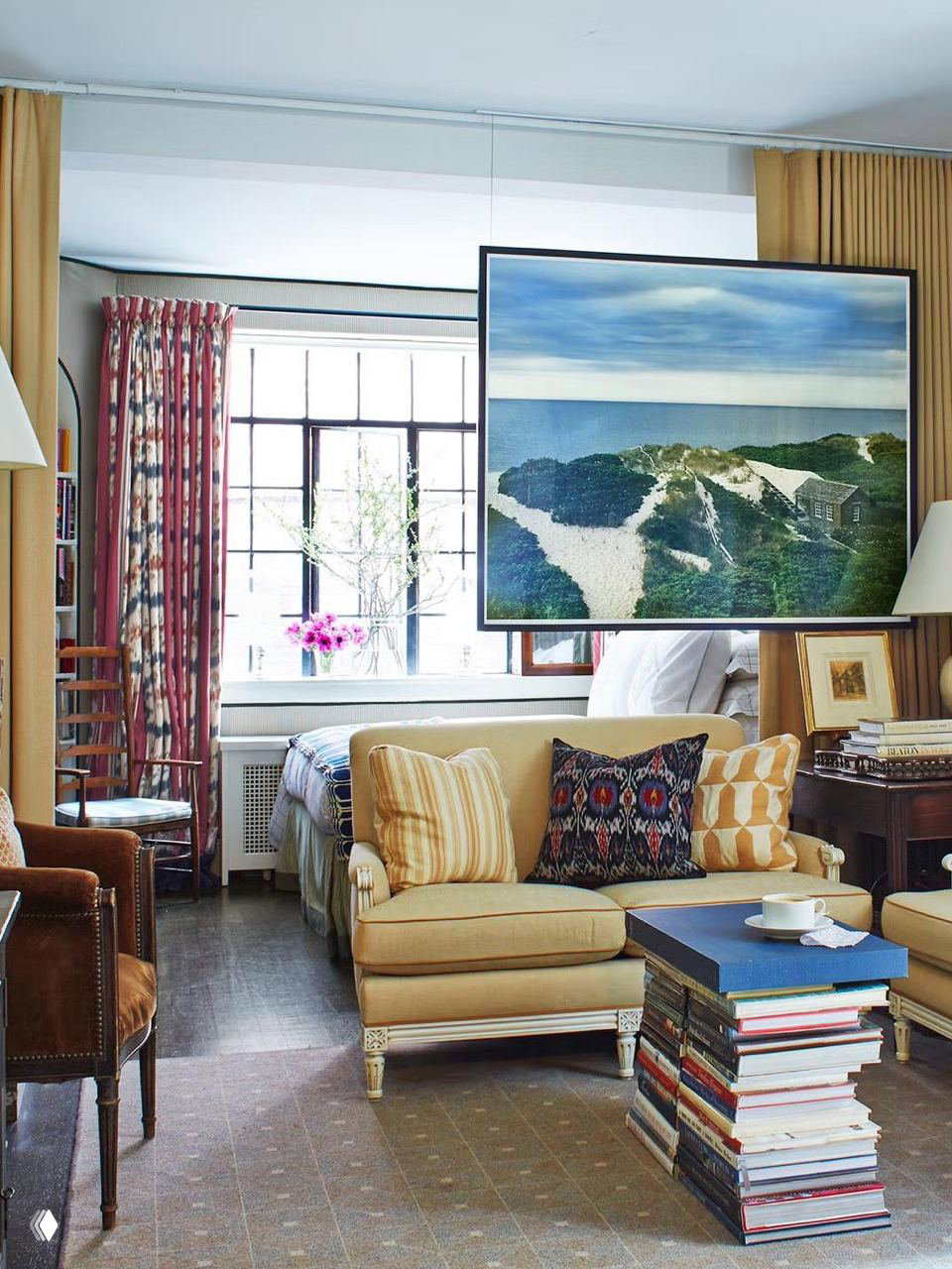 Компактная студия с подвесным телевизором над диваном, небольшим креслом и столиком из стопки книг — пример плотного совмещения гостиной и спальни в одном объёме.