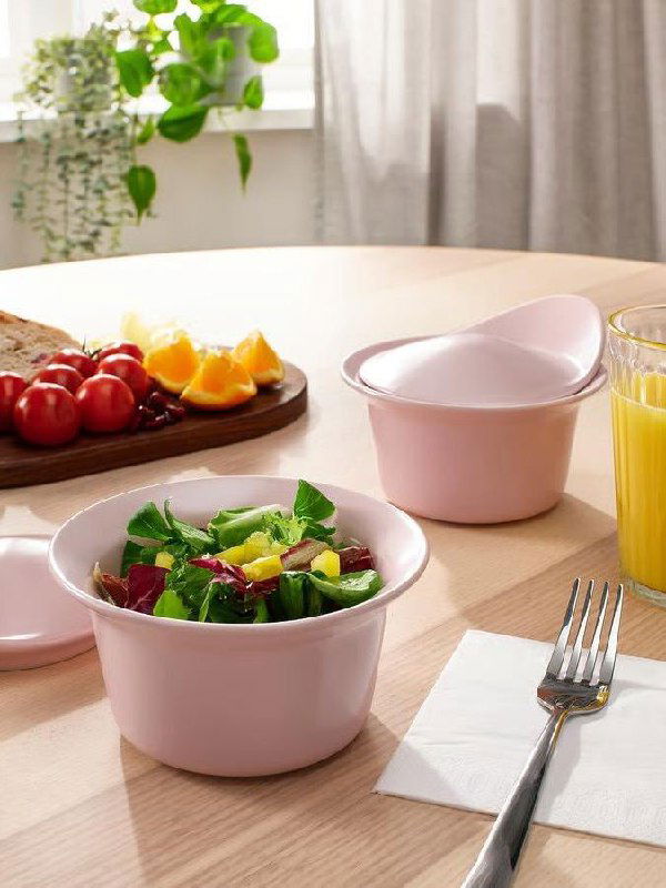 Кухонная сцена с розовыми мисками и контейнерами на деревянном столе, рядом салат в миске и стакан сока — пастельная сервировка в Rebel Pink.