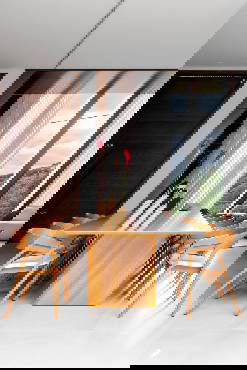 Столовая с длинным деревянным столом и стульями в едином тёплом тоне, большие окна на заднем плане обеспечивают естественный свет.