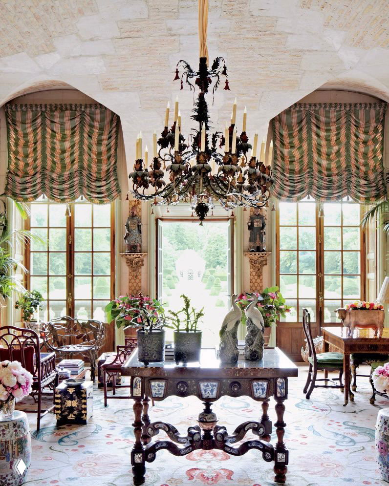 Просторная комната с высокими окнами и центральным резным столом на ковре с восточными мотивами, крупная люстра и вид на сад через двери-остекления.