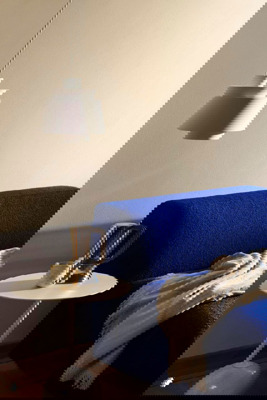 Интерьерный кадр с подвесным абажуром над круглым столиком, глубокий синий диван и мягкое покрывало, тёплый мягкий свет подчёркивает текстуры