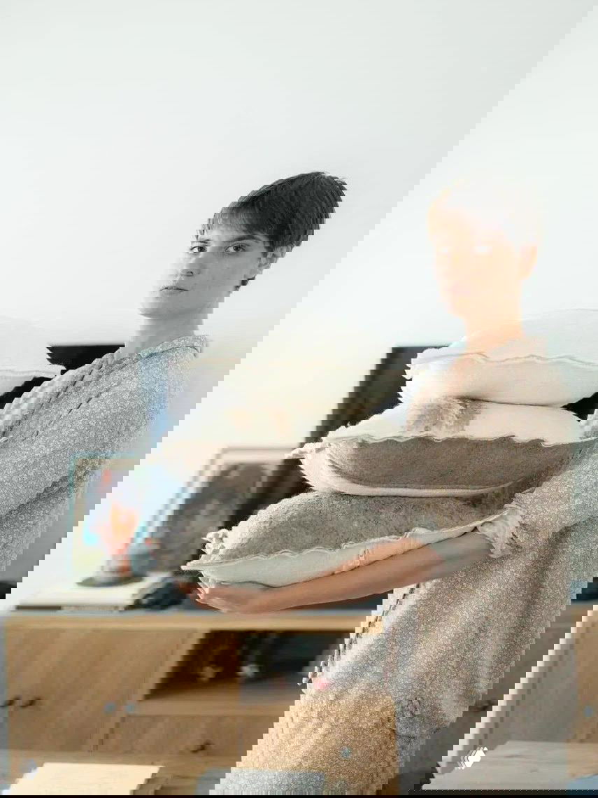 Художница держит стопку валяных подушек естественных оттенков, показывая мягкость, объем и нейтральную палитру текстильных изделий.