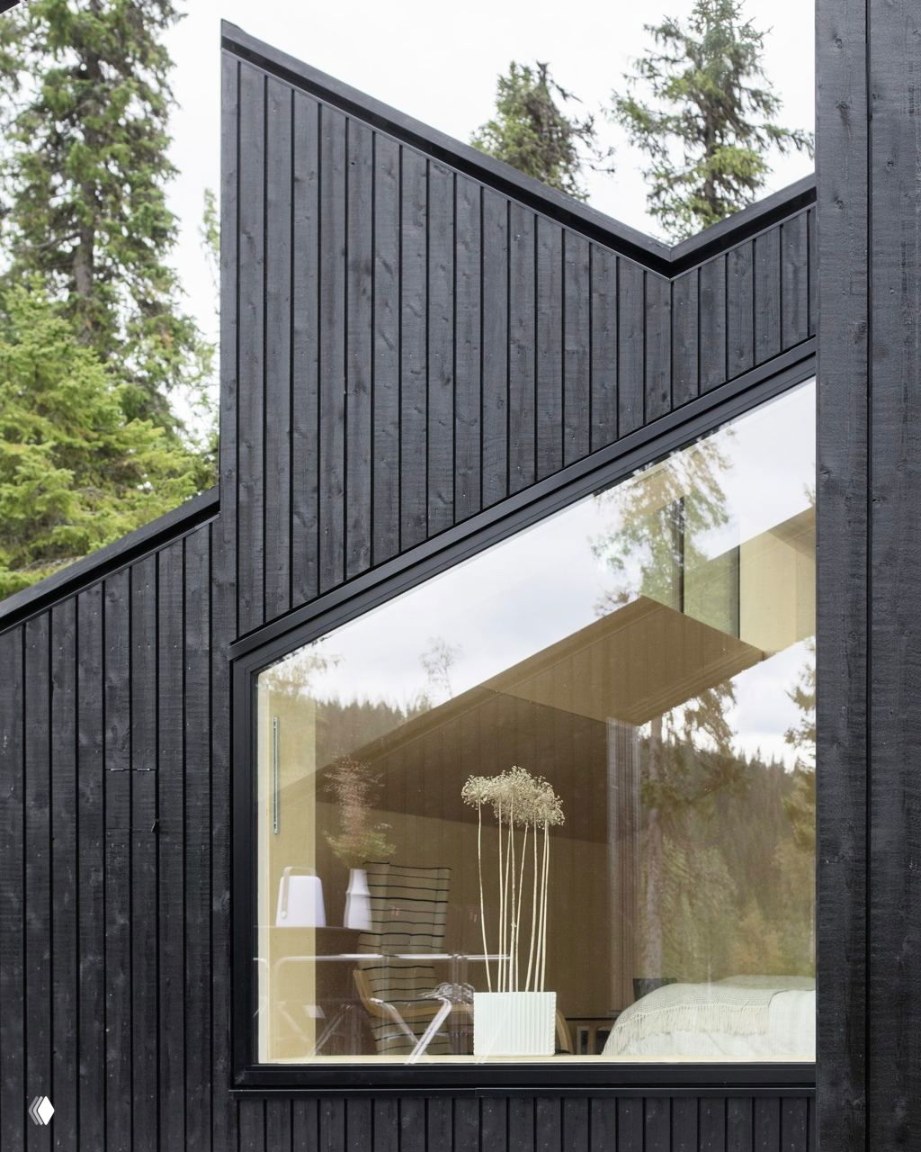 Фрагмент фасада Vindheim с крупным окном: черная деревянная обшивка и отражение хвойного леса в стекле, вид на интерьер с растением у окна.