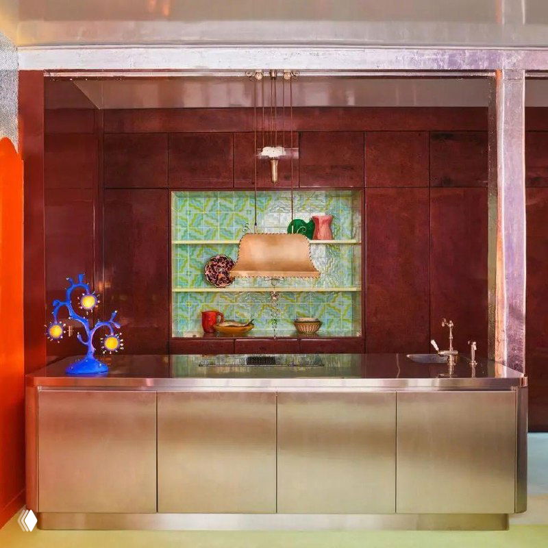 Современная кухня с металлической фасадной поверхностью, ретро-лампа и яркие декоративные предметы на фоне тёплой палитры и глянцевых деталей.