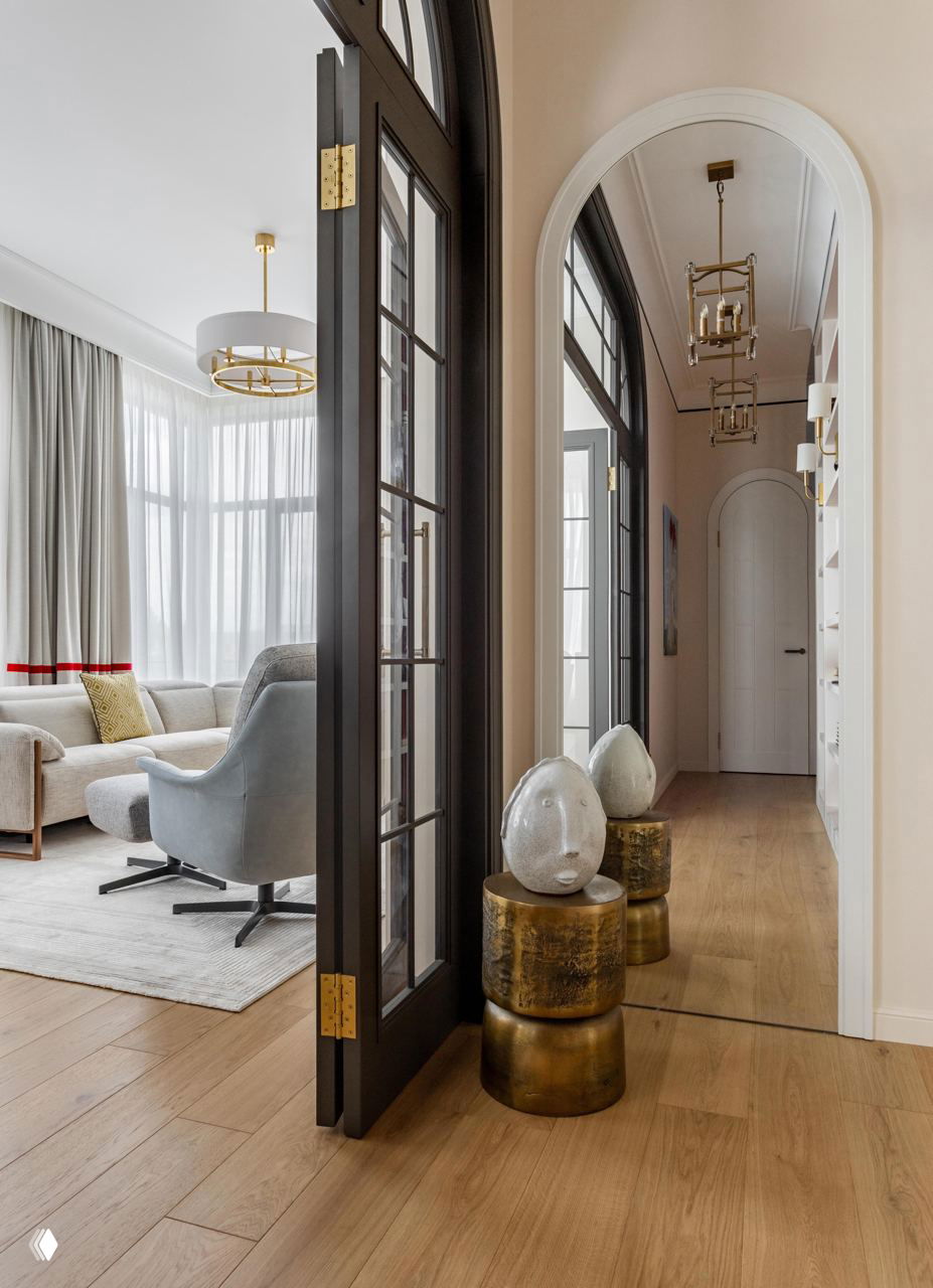 Коридор и арочные двери с зеркалом, декоративные бронзовые столики с скульптурными элементами и переход в светлую гостиную — интерьерная фотография.